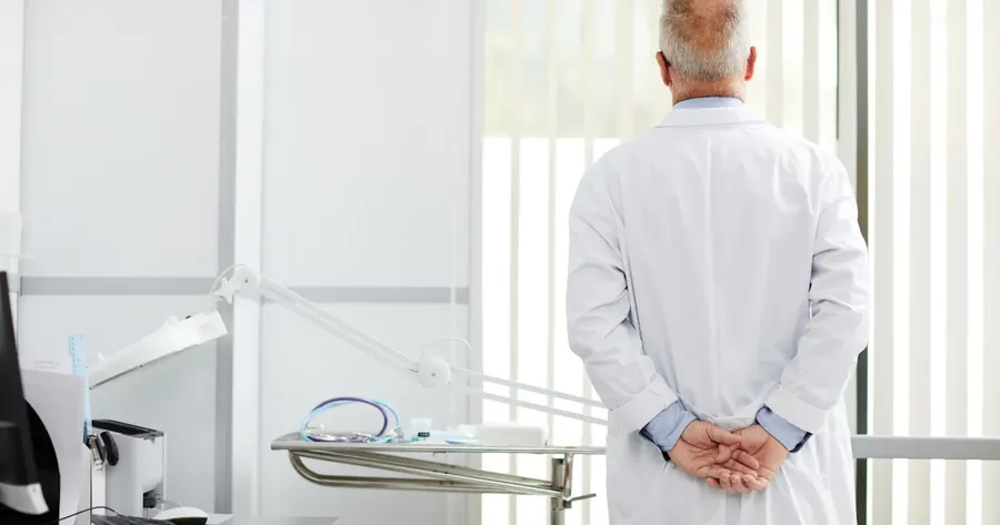 Ηλικιωμένος γιατρός με λευκή μπλούζα κοιτά έξω από το παράθυρο του άδειου ιατρείου — Ιστορίες ασφάλισης